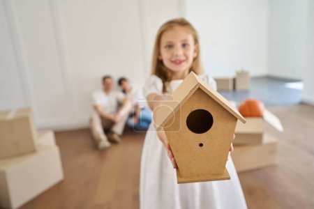 Photo for Selective focus of wooden birdhouse in hands of blurred smiling little girl and her parents on background in new modern blurred townhouse during moving. Family future planning - Royalty Free Image