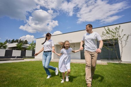 Photo for Smiling caucasian mother, father and little daughter running on green yard near modern townhouse in warm sunny day. Happy family relationship and enjoying time together - Royalty Free Image