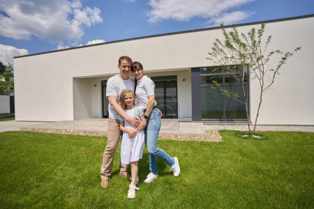 Photo for Smiling caucasian mother, father and little daughter hugging and looking at camera on green yard near modern townhouse in warm sunny day. Family relationship and spending time together - Royalty Free Image