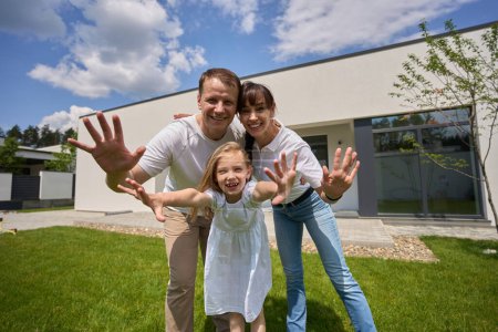 Photo for Joyful caucasian mother, father and little daughter hugging, waving hands and looking at camera on green yard near modern townhouse in warm sunny day. Family relationship and enjoying time together - Royalty Free Image