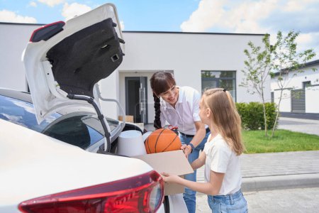 Photo for Mother and little daughter taking cardboard box with different objects from car trunk and looking at each other during moving to new modern townhouse. Family future planning and spending time together - Royalty Free Image