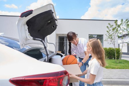 Photo for Smiling mother and daughter taking cardboard box with variety objects from car trunk and looking at each other during moving to new modern townhouse. Family future planning and spending time together - Royalty Free Image