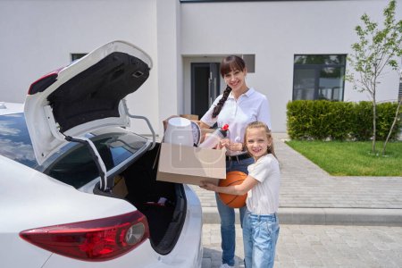 Photo for Front view of mother and daughter holding different objects from car trunk and looking at camera during moving to new modern townhouse. Family future planning, relationship and spending time together - Royalty Free Image