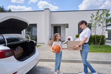Photo for Smiling mother with cardboard box and daughter with basketball ball looking at each other during moving to new modern townhouse. Family future planning, relationship and spending time together - Royalty Free Image