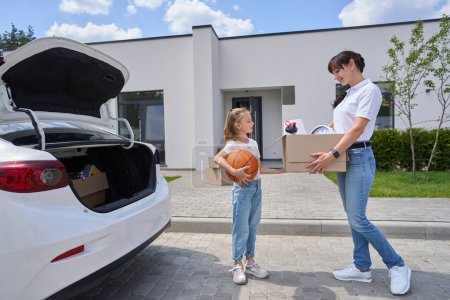Photo for Side view of mother with cardboard box and daughter with basketball ball during moving to new modern townhouse. Family future planning, relationship and spending time together - Royalty Free Image