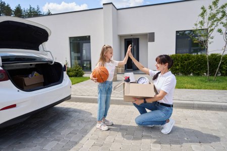 Photo for Mother with cardboard box and daughter with basketball ball giving five each other during moving to new modern townhouse. Family future planning, relationship and spending time together - Royalty Free Image