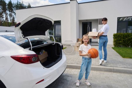 Photo for Little girl holding basketball ball while her mother carrying cardboard box with variety objects to new townhouse during moving and they looking at camera. Family future planning - Royalty Free Image