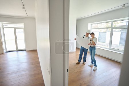 Photo for Focused caucasian male worker and woman watching blueprint in new modern comfortable townhouse before repair - Royalty Free Image