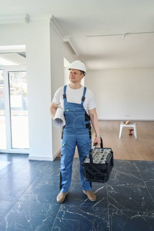 Foto de Sonriente adulto caucásico trabajador masculino con plano y caja de herramientas mirando hacia otro lado en la nueva casa moderna y cómoda antes de la reparación - Imagen libre de derechos