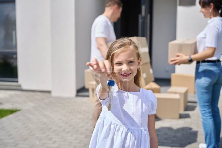 Téléchargez les photos : Fille dans la cour avec les clés d'une nouvelle maison, adultes portant des boîtes de choses dans la maison - en image libre de droit