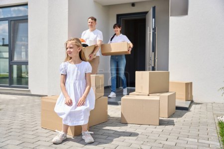 Téléchargez les photos : Une fille en robe blanche est assise sur une boîte dans la cour, ses parents portent les boîtes dans la maison - en image libre de droit