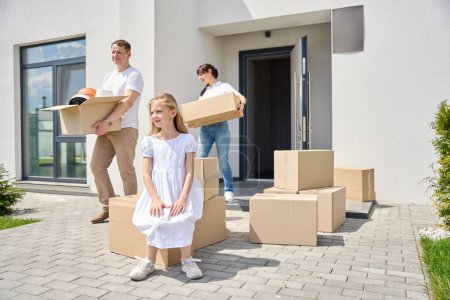 Photo for Girl sits on a box in the yard, her parents carry boxes of things - Royalty Free Image