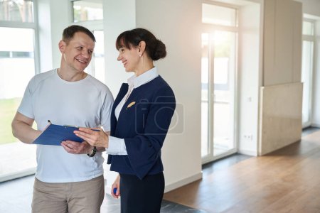 Téléchargez les photos : Acheteur satisfait signe des documents pour l'achat d'une maison, à côté d'une femme souriante agent immobilier - en image libre de droit