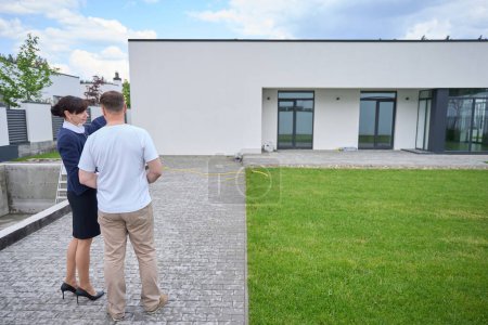 Verkäuferin mit einem Kunden bei der Besichtigung eines neuen Hauses, im Hof befindet sich ein Swimmingpool