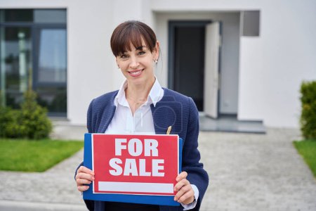 Junge Frau steht in der Nähe eines Landhauses mit einem Verkaufsschild, ein Makler im Business-Anzug