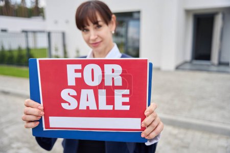 Téléchargez les photos : Femme agent immobilier se tient avec un signe à vendre, une dame dans un costume d'affaires - en image libre de droit