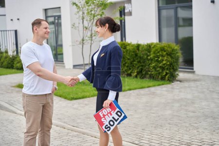 Immobilienmaklerin begrüßt einen Käufer an einer neuen Hütte, sie hält ein zum Verkauf stehendes, verkauftes Schild in ihren Händen