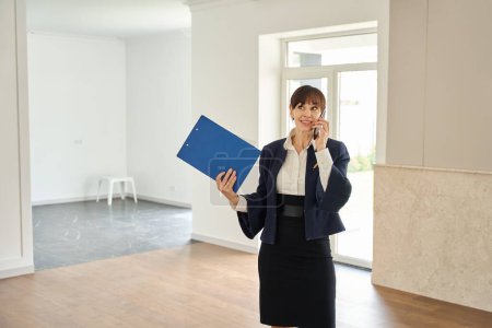 Photo for Female sales manager communicates on her mobile phone in a spacious room, she has a blue folder with documents - Royalty Free Image