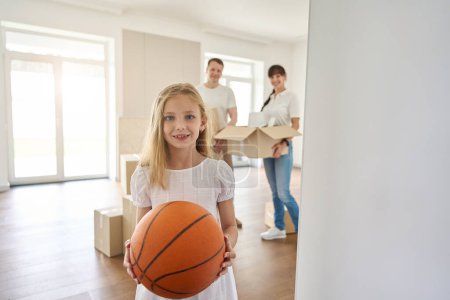 Foto de Chica con una pelota se para en una habitación espaciosa, al lado de sus padres están llevando cajas de cosas - Imagen libre de derechos