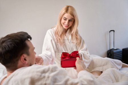 Téléchargez les photos : Heureux jeunes mariés sont assis dans des peignoirs confortables sur le lit, un homme remet une boîte cadeau à une femme - en image libre de droit
