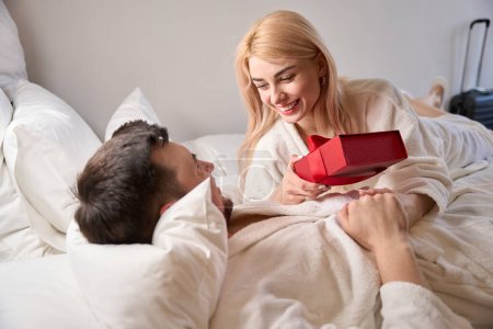 Joyful newlyweds are seated in cozy bathrobes on the bed, the woman is holding a gift box in her hands
