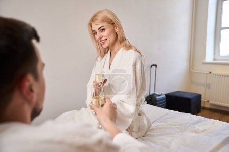 Téléchargez les photos : Les jeunes mariés avec une coupe de champagne sont assis sur un grand lit, avec des bagages de voyage à proximité - en image libre de droit