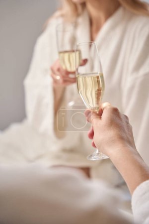 Téléchargez les photos : Couple romantique avec une coupe de champagne assis sur un grand lit, les gens en peignoirs moelleux - en image libre de droit