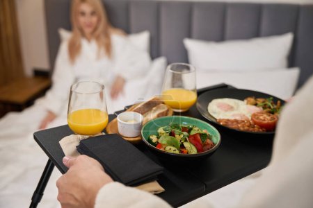 Téléchargez les photos : Jeune mari porte un plateau de petit déjeuner au lit, le repas est servi pour deux - en image libre de droit