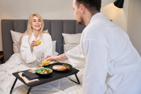 Foto de Joven marido trajo una bandeja de desayuno a su esposa en la cama, los recién casados se relajan en una habitación cómoda - Imagen libre de derechos