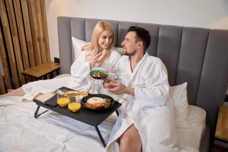 Foto de Los recién casados están desayunando juntos en una cómoda habitación de hotel, están ubicados en un acogedor dormitorio - Imagen libre de derechos