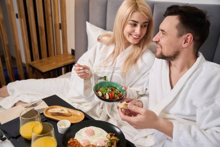 Photo for Young blonde woman and her husband are having breakfast in comfortable hotel bedroom, they are accommodated in a honeymoon suite - Royalty Free Image