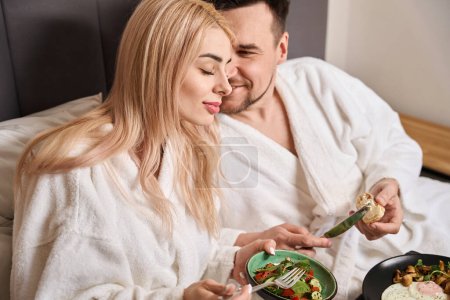 Téléchargez les photos : Les jeunes conjoints prennent le petit déjeuner dans une chambre d'hôtel confortable, ils sont hébergés dans une suite nuptiale - en image libre de droit