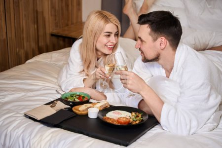 Téléchargez les photos : Les jeunes mariés apprécient le petit déjeuner au lit avec du champagne, ils sont dans des peignoirs moelleux - en image libre de droit