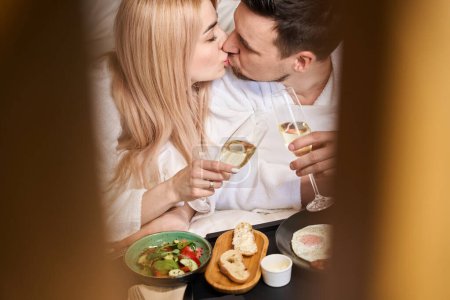Téléchargez les photos : Heureux jeunes mariés profiter du petit déjeuner au lit avec du champagne, ils embrassent tendrement - en image libre de droit