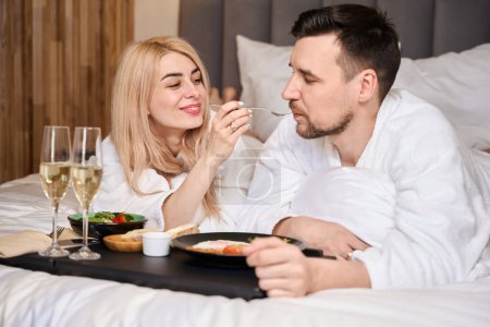 Téléchargez les photos : Jeune couple marié profitant du petit déjeuner au lit, deux verres de champagne sur un plateau - en image libre de droit