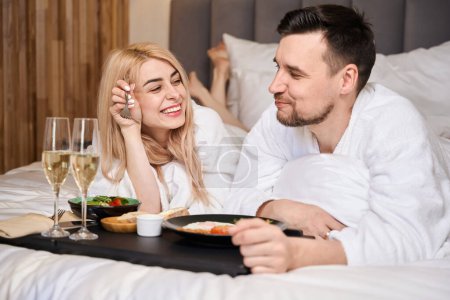 Foto de Joven pareja casada se comunica durante el desayuno en la cama, hay dos copas de champán en una bandeja - Imagen libre de derechos