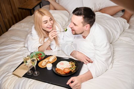 Foto de Alegre pareja joven hablando durante el desayuno en la cama, dos copas de champán en una bandeja - Imagen libre de derechos