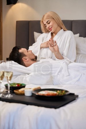 Foto de La pareja casada se relaja en una cama grande, el desayuno con champán se sirve en una bandeja - Imagen libre de derechos