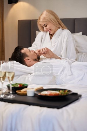 Téléchargez les photos : Homme et femme se prélassent sur un grand lit dans une chambre d'hôtel, le petit déjeuner avec champagne est servi sur un plateau - en image libre de droit