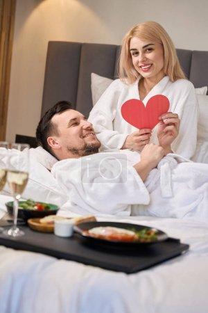 Téléchargez les photos : Les jeunes mariés se prélassent sur le lit dans leur chambre d'hôtel, prennent le petit déjeuner avec du champagne et un cœur cadeau mignon - en image libre de droit