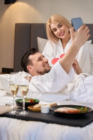 Foto de Recién casados toman selfie en la cama en una habitación de hotel, están desayunando con champán y un lindo corazón de regalo - Imagen libre de derechos