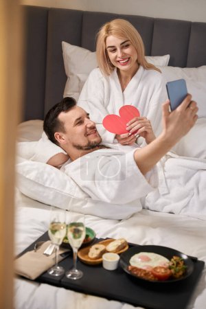Foto de Pareja toma selfie en la cama en la habitación del hotel, desayunan con champán y un lindo corazón de regalo - Imagen libre de derechos