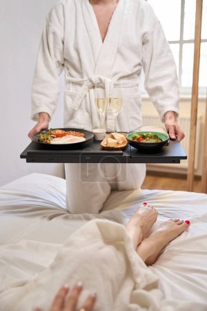 Téléchargez les photos : Le mari a apporté le petit déjeuner avec du champagne à sa jeune femme au lit, le couple dans la chambre des jeunes mariés - en image libre de droit