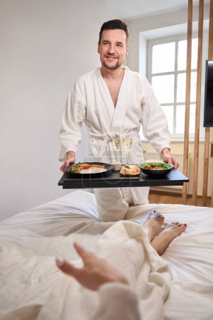 Foto de El hombre trajo el desayuno con champán a su joven esposa en la cama, una pareja en una habitación recién casada - Imagen libre de derechos