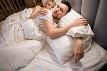 Téléchargez les photos : L'homme éveillé repose sur un lit confortable, sa jeune femme sommeille à côté de lui - en image libre de droit