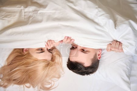 Téléchargez les photos : Joyeux époux confortablement nichés sous une couverture sur un grand lit, chambre de couple pour les jeunes mariés - en image libre de droit