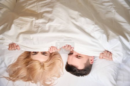 Foto de Hombre y una mujer se sientan cómodamente debajo de una manta en una cama grande, una habitación de parejas para recién casados - Imagen libre de derechos