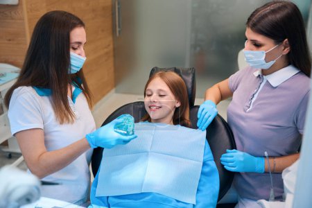 Child is having a consultation with a dental hygienist in a modern clinic, the doctor is working with an assistant