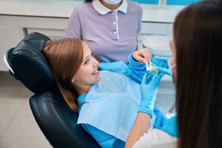 Foto de Dentista mujer demuestra un modelo de una dentición a una paciente chica, con un asistente dental cerca - Imagen libre de derechos