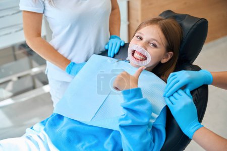 Téléchargez les photos : Enfant montre ok à un rendez-vous chez le dentiste, la fille a un rétracteur dentaire dans la bouche - en image libre de droit